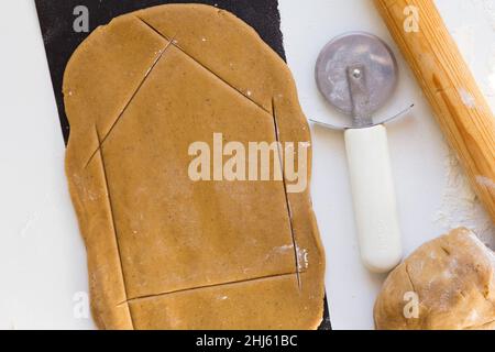 Cutting out wall of gingerbread house with knife for pizza on rolled raw gingerbread dough teflon baking sheet on the white background Stock Photo