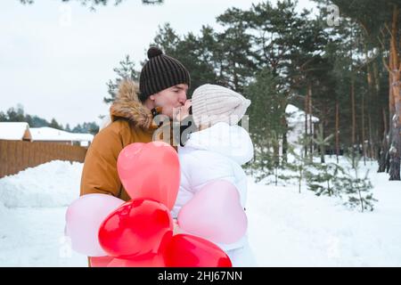 A man and a woman in love kissing on outdoor date in winter in the snow with a gift of pink and red balloons in the shape of a heart. Valentine's Day, Stock Photo