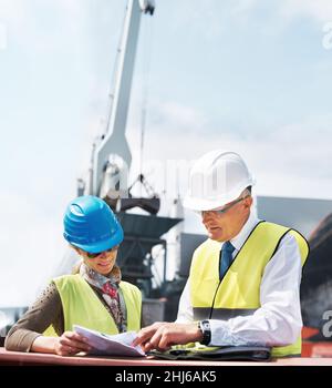 Working together can strengthen any foundation. Two dock workers holding paperwork while standing in the shipyard. Stock Photo