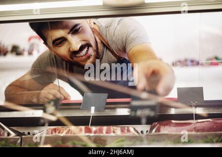 Young man butcher arranging meat products in display case of butcher shop Stock Photo