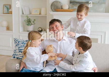 An annoyed father sitting on sofa at home in living room distracted from noise and touching his temple, hyperactive little children attract attention of children who want to play. Tired dad Stock Photo