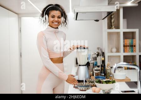 Black woman in headphones smiling and making smoothie at home Stock Photo