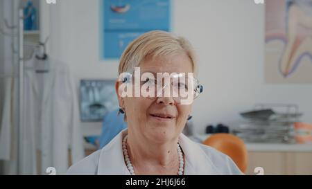 Portrait of senior dentist sitting in stomatology office, wearing protective glasses. Stomatologist preparing for dentistry appointment, working in oral care cabinet. Close up Stock Photo