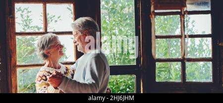 Senior couple in love dance and enjoy happy life together at home with outdoor nature in background outside the window. Mature elderly people enjoy re Stock Photo