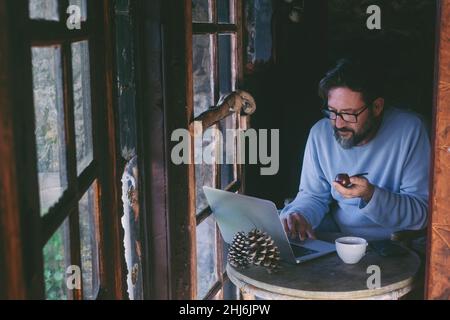 Mature man sitting at home working on laptop computer and smoking pipe. Alternative single lifestyle male people indoor in cozy cabin wooden chalet. T Stock Photo