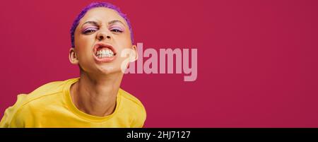 Quirky young woman making a face in a studio. Fashionable young woman looking at the camera confidently while standing against a pink background. Styl Stock Photo
