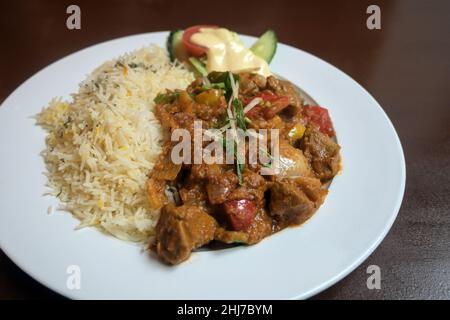 Indian curry made from meat and vegetables in a spicy sauce served with rice and salad decoration on a white plate and a brown wooden table, selected Stock Photo