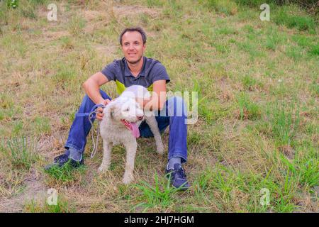 man with a white dog of a laggoto-Romagnolo breed sitting on grass in the park. Walking the dog in the park. dog in search of truffle mushrooms Stock Photo