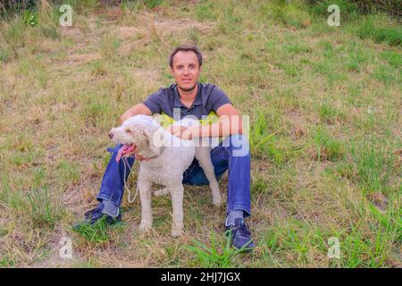 man with a white dog of a laggoto-Romagnolo breed sitting on grass in the park. Walking the dog in the park. dog in search of truffle mushrooms Stock Photo