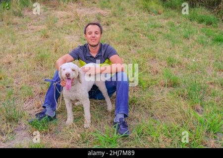 man with a white dog of a laggoto-Romagnolo breed sitting on grass in the park. Walking the dog in the park. dog in search of truffle mushrooms Stock Photo