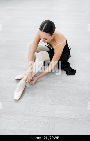 young ballerina adjusting pointe shoe while sitting on floor in studio Stock Photo