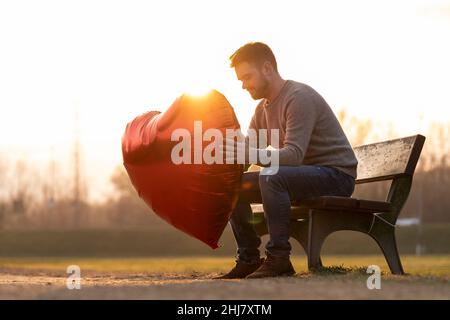 Sad young man holding up a heart-shaped balloon sitting on a bench in the park Stock Photo