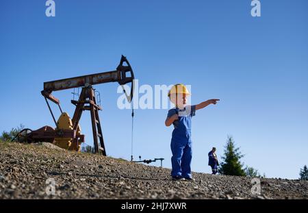 Adorable child pointing finger at copy space while standing in oil field with petroleum pump jack and blue sky on background. Snapshot of oil filed with cute little boy and oil pump rocker-machine. Stock Photo