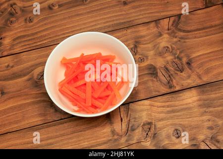 White bowl filled with Carrot sticks placed on a Oak coloured wood board background Stock Photo