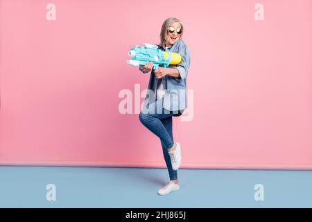 Full length photo of funny aged lady go play water gun wear jeans shirt isolated on pink background Stock Photo