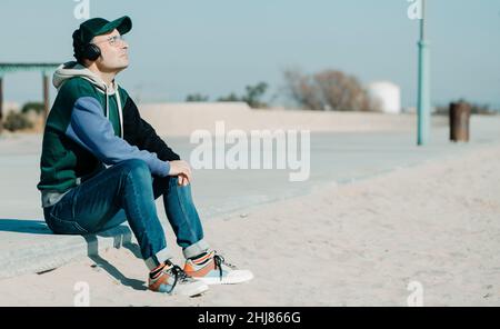 a middle-aged man, wearing a multicolored hoodie and jeans, listens to music using a pair of headphones sitting on the promenade, next to the sand of Stock Photo