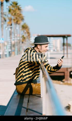 closeup of a mature man, wearing colorful modern casual clothes, watching his smartphone, sitting outdoors next to the sand of a urban beach Stock Photo