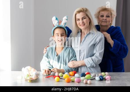 Smiling happy Caucasian grandmother, mother and daughter painting and decorating Easter eggs having great time together in home kitchen Stock Photo