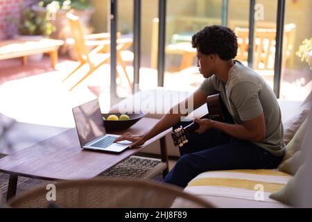 Side view of african american young man using laptop while sitting with guitar at home. unaltered, music, wireless technology, food, learning, lifesty Stock Photo