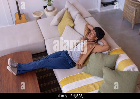 African american young man relaxing on sofa with hands behind head and feet up on table at home. unaltered, lifestyle, relaxation, leisure activity an Stock Photo