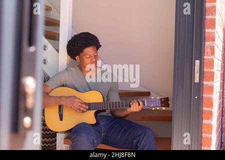 African american young man playing guitar while sitting on steps at home. unaltered, music, lifestyle, leisure activity and domestic life concept. Stock Photo