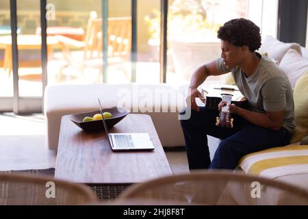 African american young man holding guitar while looking in laptop at table. unaltered, music, wireless technology, learning, lifestyle, food, leisure Stock Photo