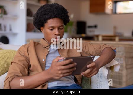 African american young man using digital tablet while sitting on sofa at home. unaltered, lifestyle, leisure activity, wireless technology, concentrat Stock Photo
