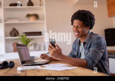Smiling african american young businessman using smart phone while working on laptop at home. unaltered, business, wireless technology, reading, data Stock Photo