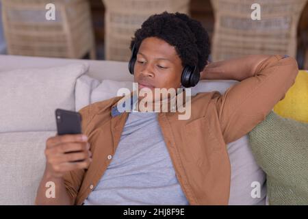 African american young man using smart phone while listening music on headphones at home. unaltered, lifestyle, leisure activity, wireless technology, Stock Photo