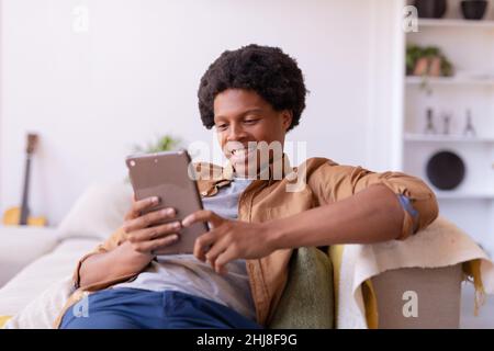 Smiling african american young man using digital tablet while sitting on sofa at home. unaltered, lifestyle, leisure activity, wireless technology, wa Stock Photo