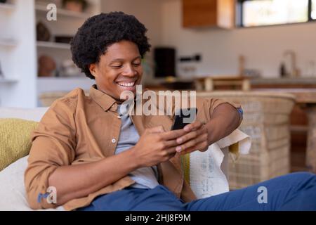 Cheerful african american young man using smart phone while sitting on sofa at home. unaltered, lifestyle, leisure activity, wireless technology, soci Stock Photo