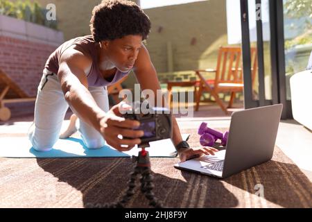 African american young man adjusting camera while using laptop for making vlog on exercise at home. unaltered, fitness, vlogging, influencer, wireless Stock Photo