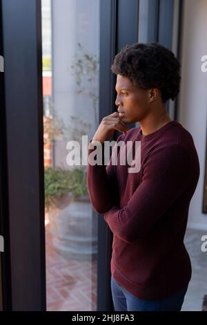 Thoughtful african american young man with hand on chin standing by window at home. unaltered, lifestyle, contemplation, day dreaming and domestic lif Stock Photo