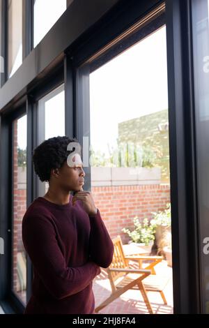 Thoughtful african american young man with hand on chin looking through window at home. unaltered, lifestyle, contemplation, day dreaming and domestic Stock Photo