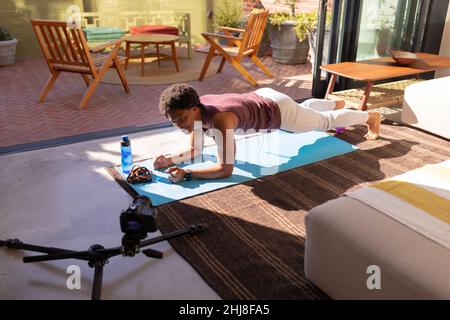 Smiling african american young man doing push-ups on exercise mat while making a vlog with camera. unaltered, fitness, vlogging, influencer and active Stock Photo