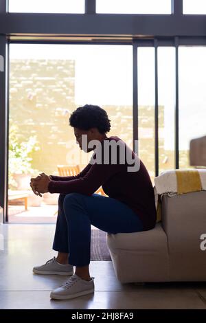 Side view of thoughtful african american young man with hands clasped sitting on sofa at home. unaltered, lifestyle, contemplation, day dreaming, depr Stock Photo