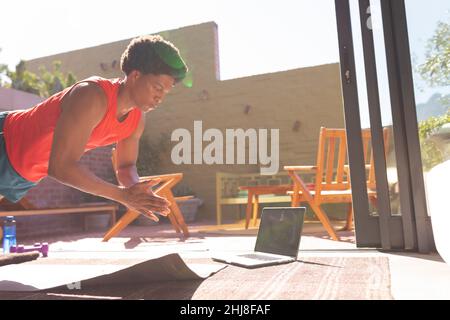 African american young man doing clapping push ups on exercise mat by laptop at home. unaltered, fitness, wireless technology, influencer, balance and Stock Photo