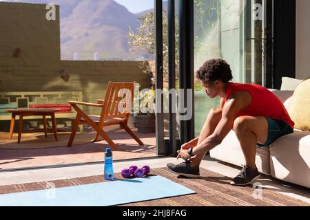 African american young man tying shoes lace while sitting on sofa at home. unaltered, fitness and active lifestyle concept. Stock Photo