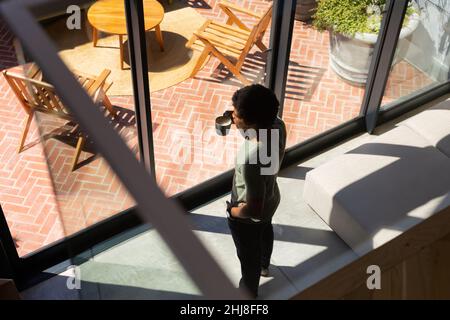 High angle view of african american young man with coffee cup standing by window at home. unaltered, lifestyle, drink, contemplation, day dreaming and Stock Photo