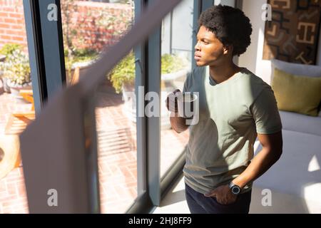 Sunlight falling on thoughtful young man with coffee cup standing by window at home. unaltered, lifestyle, drink, contemplation, day dreaming and dome Stock Photo