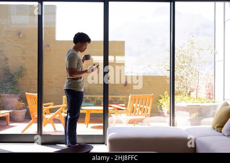 Full length of african american young man using smart phone while standing with coffee cup by window. unaltered, lifestyle, leisure activity, drink, s Stock Photo