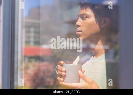 Close-up of thoughtful african american young man with coffee cup looking through window. unaltered, lifestyle, day dreaming, contemplation and domest Stock Photo