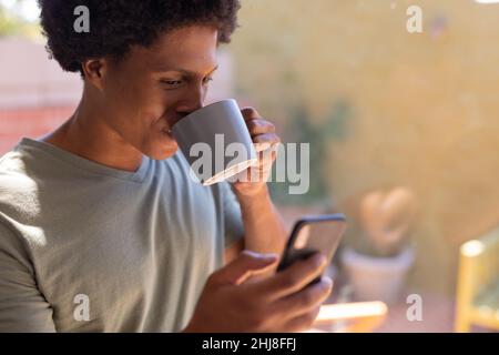 Close-up of smiling african american young man drinking coffee while using smart phone at home. unaltered, lifestyle, leisure activity, drink, social Stock Photo
