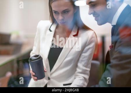 Two colleagues discuss project updates while having a coffee break. Young business people in the suits with coffee in hands looking down standing behind the glass.  Stock Photo