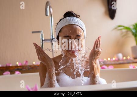 Smiling biracial young woman washing face in bathtub at spa. unaltered, spa, body care, lifestyle, vitiligo and wellness. Stock Photo