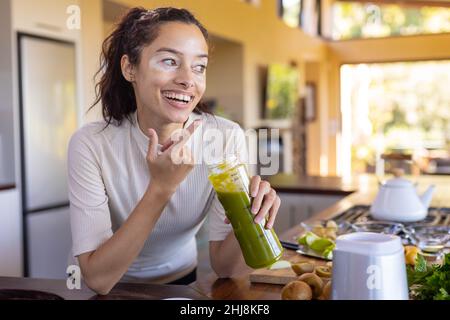 Cheerful biracial young woman pointing and looking away while having fruit juice in kitchen at home. unaltered, lifestyle, vitiligo and healthy food c Stock Photo