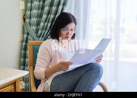 Serious mature woman reading papers documents sitting in armchair at home. Stock Photo