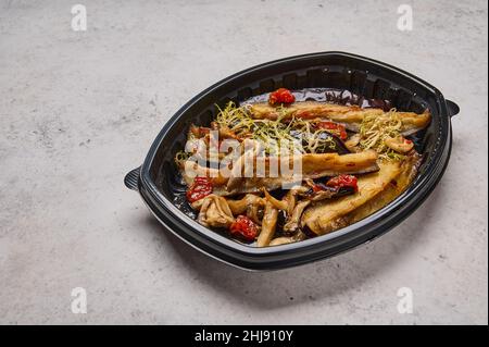 Crispy eggplant with baked cherry tomatoes and oyster mushrooms in black plastic container on light background. Designer food concept for take away or delivery Stock Photo