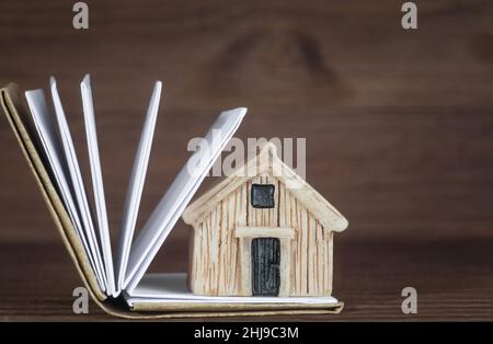 Close-up of a miniature house model placed on a small open book with blank pages against a wooden background. Creating a household manual. Stock Photo