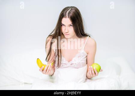 The girl sits on the bed, wrapped in a blanket, and holds a banana and an apple, chooses what to eat. The brunette is having breakfast with fruit. Stock Photo